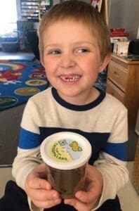 Smiling young boy with short blond hair wearing a cream sweater with blue stripes, holding a jar of soil with a lid that has a worm graphic and text on it, likely a classroom worm science project.