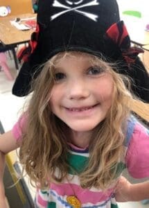 Young girl wearing a black pirate hat with a skull and crossbones, smiling at the camera in a colorful striped dress.
