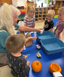 ps 1028 1 - First Lutheran Church and School, Helena, MT Teacher guiding a group of children at a blue table with orange pumpkins and craft supplies in a classroom divider bins nearby.